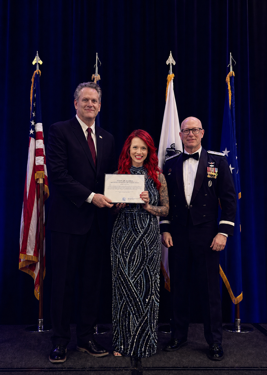 Nicole Gebhardt holding a certificate, flanked by two men in formal attire. One man wears a dark suit, and the other wears a military dress uniform with medals. They pose in front of a row of flags, including the U.S. flag, against a dark blue curtain backdrop.